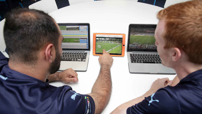 Two Sydney FC players watch soccer video footage analysis on two laptops and a tablet